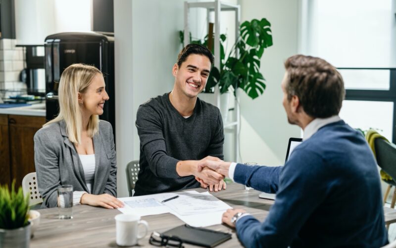 Young happy couple making an agreement with their financial advisor at home. Men are shaking hands.